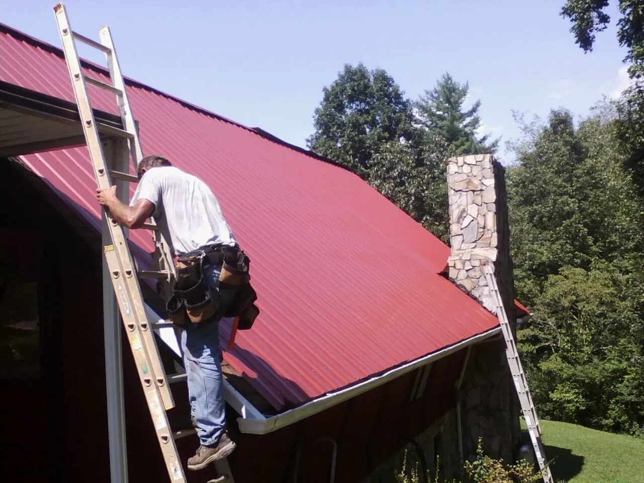 Roofer on a ladder beside a red metal roof and stone chimney — Soffit & Fascia Repair by Roofing Brothers in Delhi